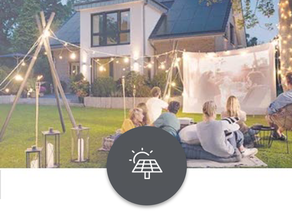 Solaranlage Eine Familie sitzt auf dem Rasen vor einem Haus mit Solaranlage und schaut einen Film auf einer Projektionsleinwand.